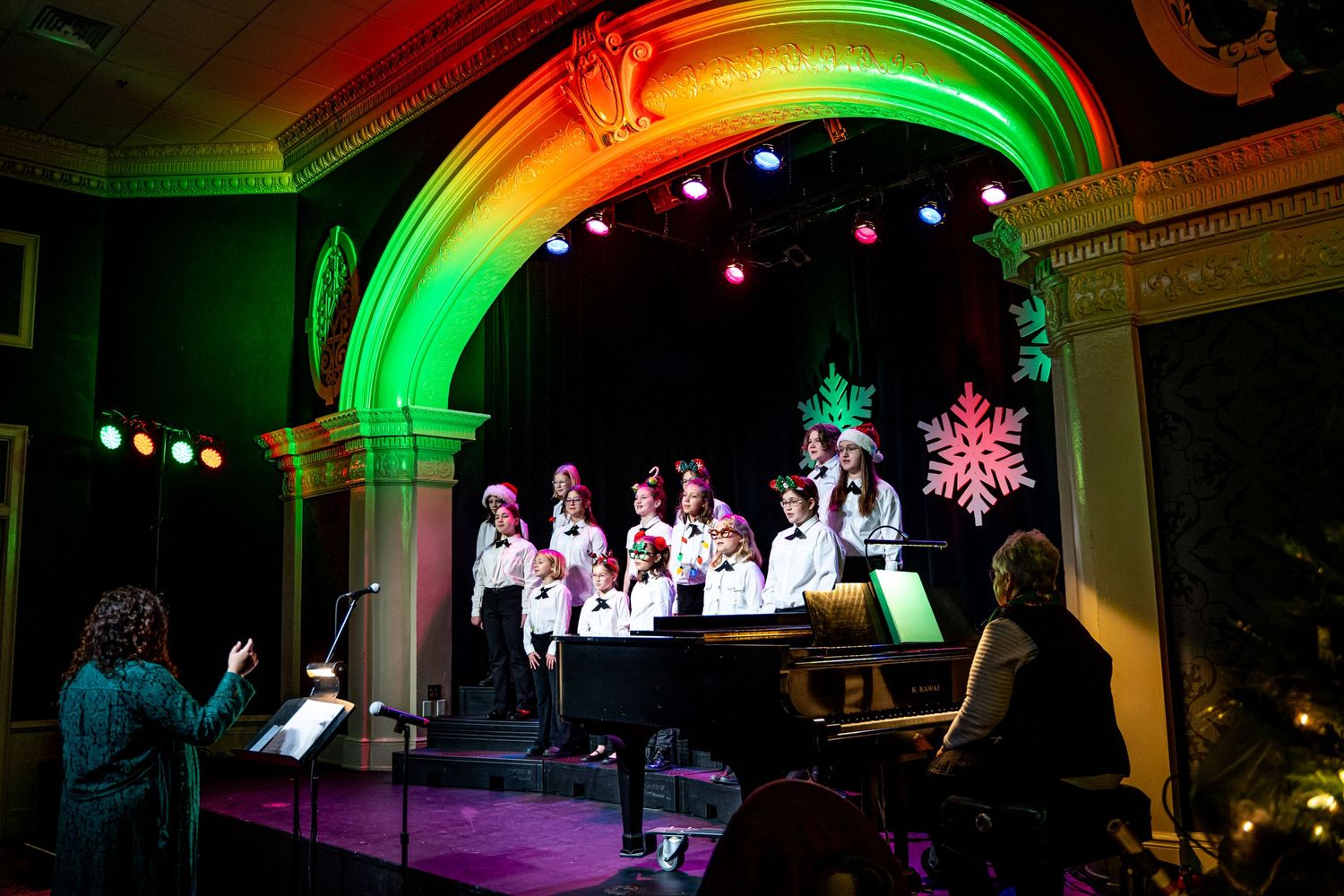 Children's choir performs on stage with colorful Christmas decorations and green arch lighting during holiday concert.