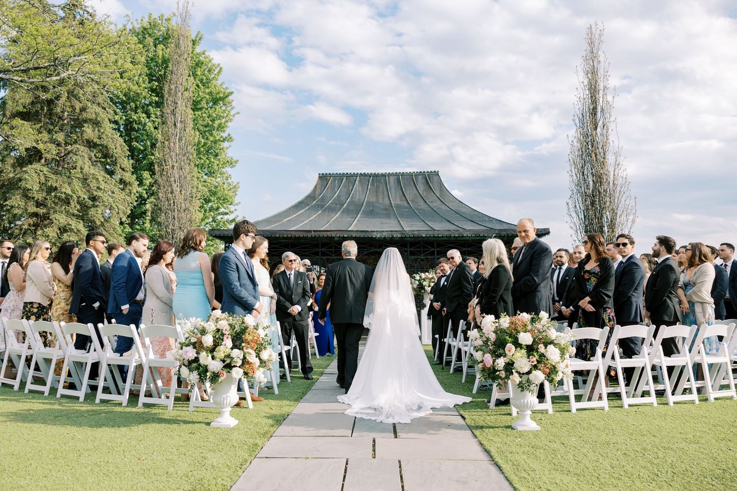 Bride walking down outdoor aisle at elegant garden wedding ceremony with white chairs and floral arrangements.