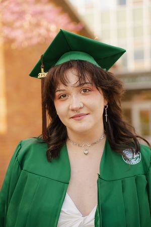 Young woman in green graduation cap and gown smiling outdoors during spring commencement ceremony.