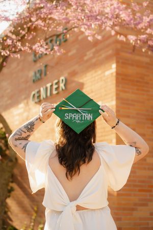 Graduate holding green Spartan mortarboard cap in front of brick Art Center building with pink cherry blossoms.