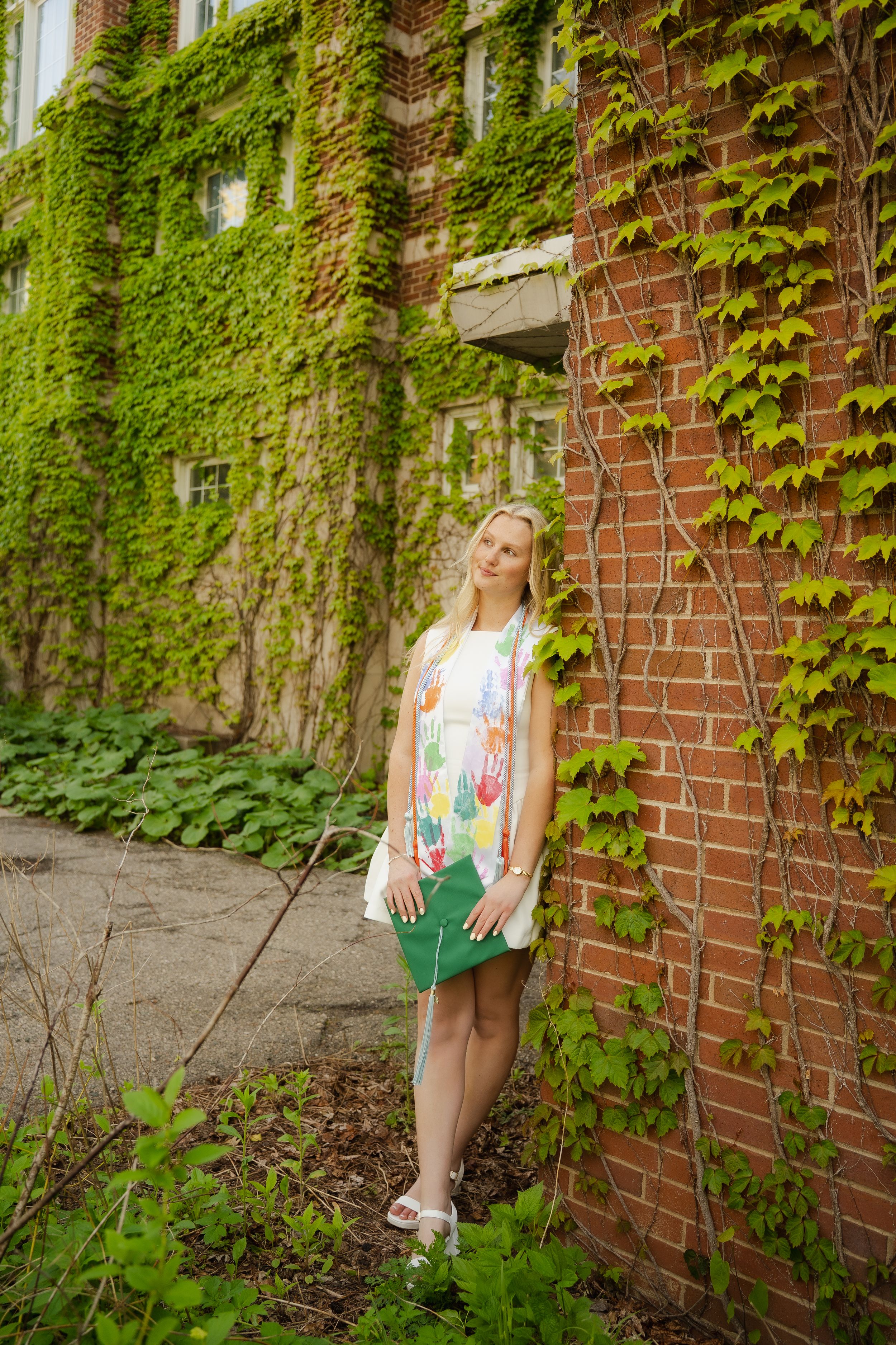 Blonde woman in floral dress posing against ivy-covered brick wall outdoors.
