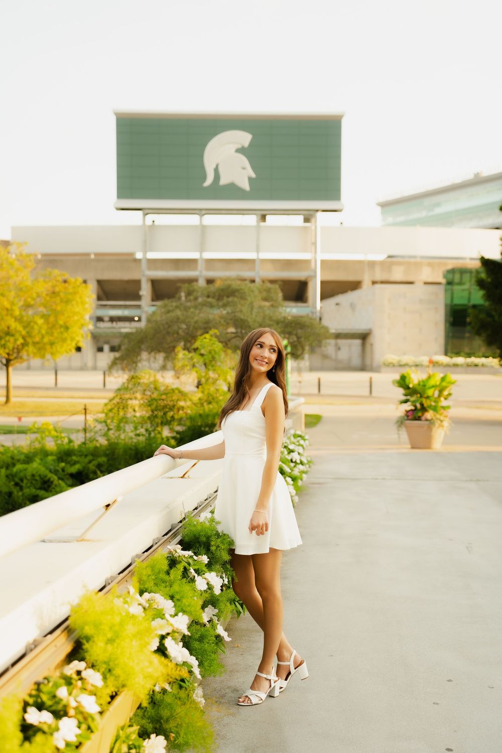 Young woman in white dress posing near flowers with Michigan State Spartan logo sign in background.