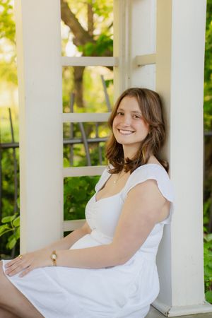 Young woman in white dress smiling while sitting on a porch between white columns with green trees behind her.