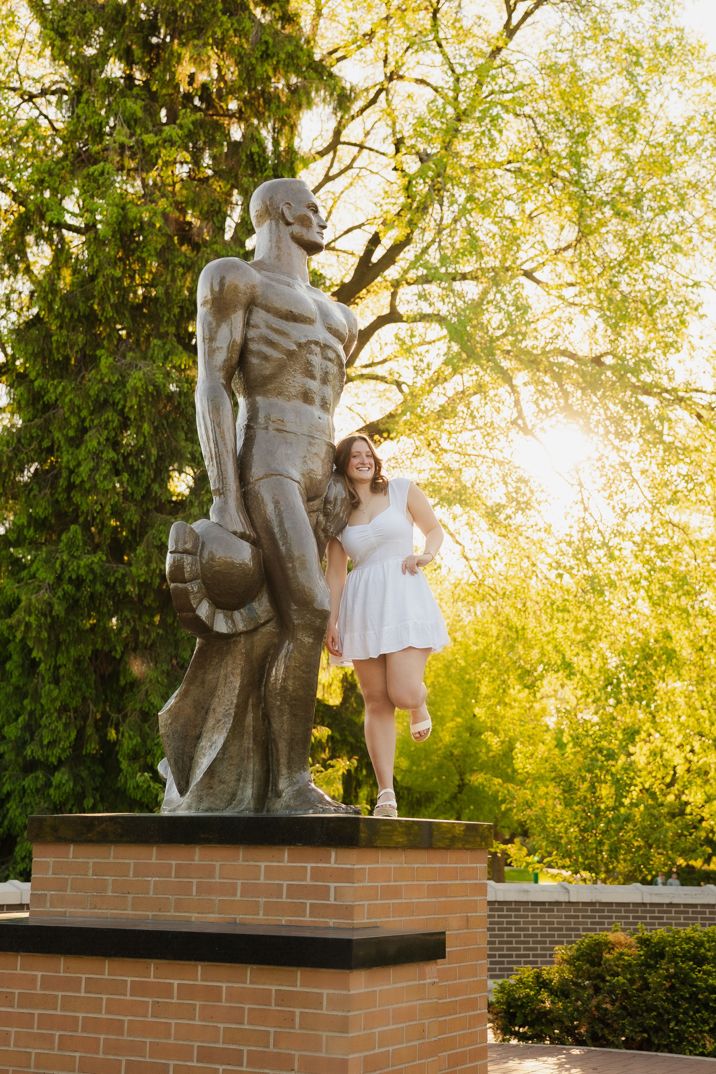 Young woman in white dress poses beside a large bronze Spartan statue on brick pedestal, surrounded by lush green trees in sunlight.