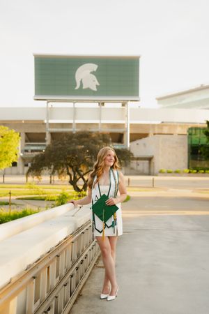 Young woman in white dress holding green clutch poses near Michigan State University Spartan Stadium sign.
