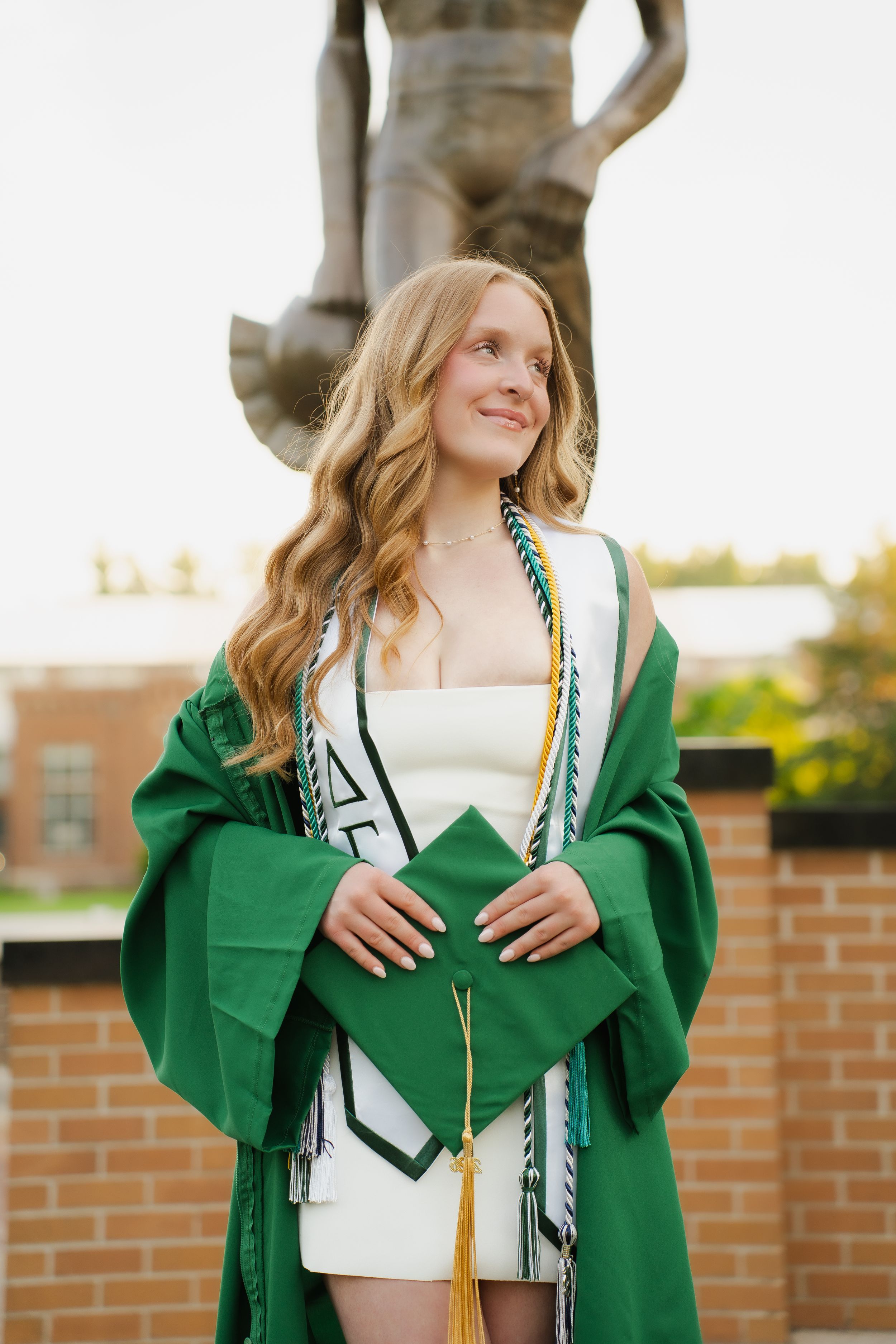 Young woman in green graduation gown and cap holding honors cords, smiling outdoors near a brick building with statue.