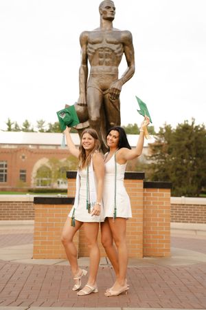 Two women in white dresses celebrate graduation holding green caps near a bronze Spartan statue at Michigan State University.
