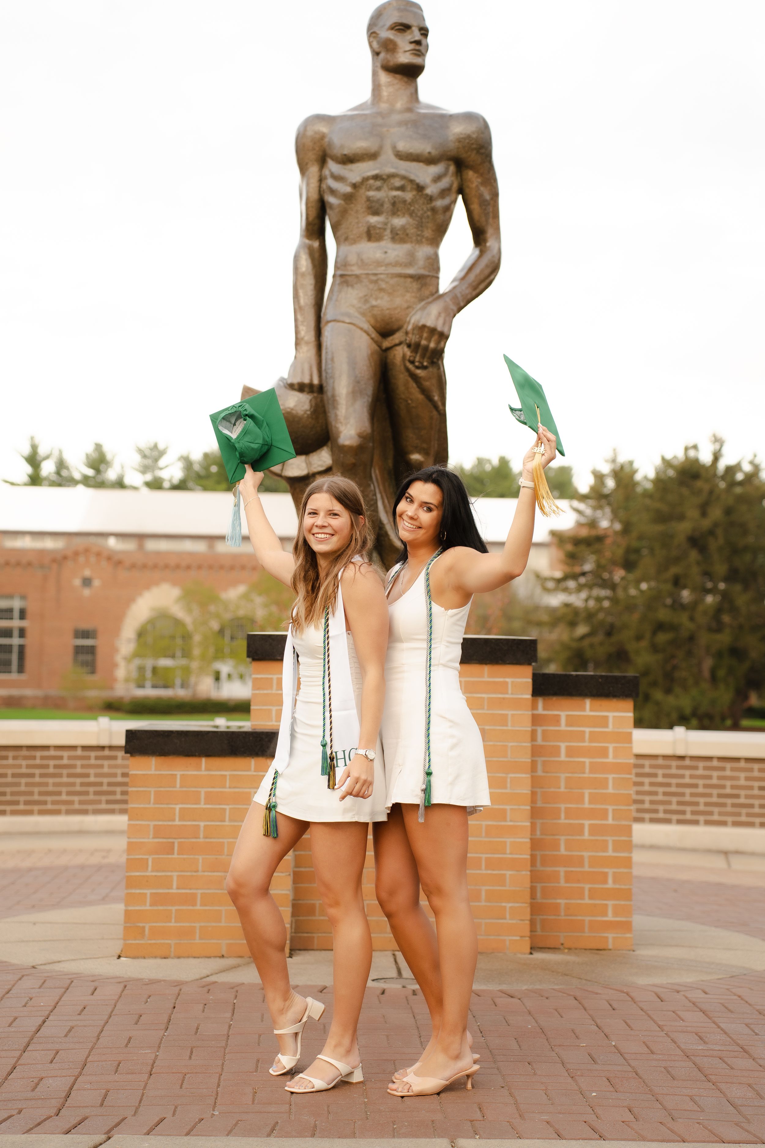 Two women in white dresses celebrate graduation holding green caps near a bronze Spartan statue at Michigan State University.