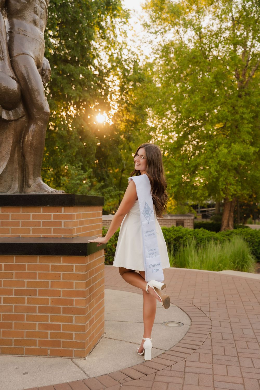 Young woman in white dress and graduation sash posing by brick statue pedestal outdoors at golden hour.