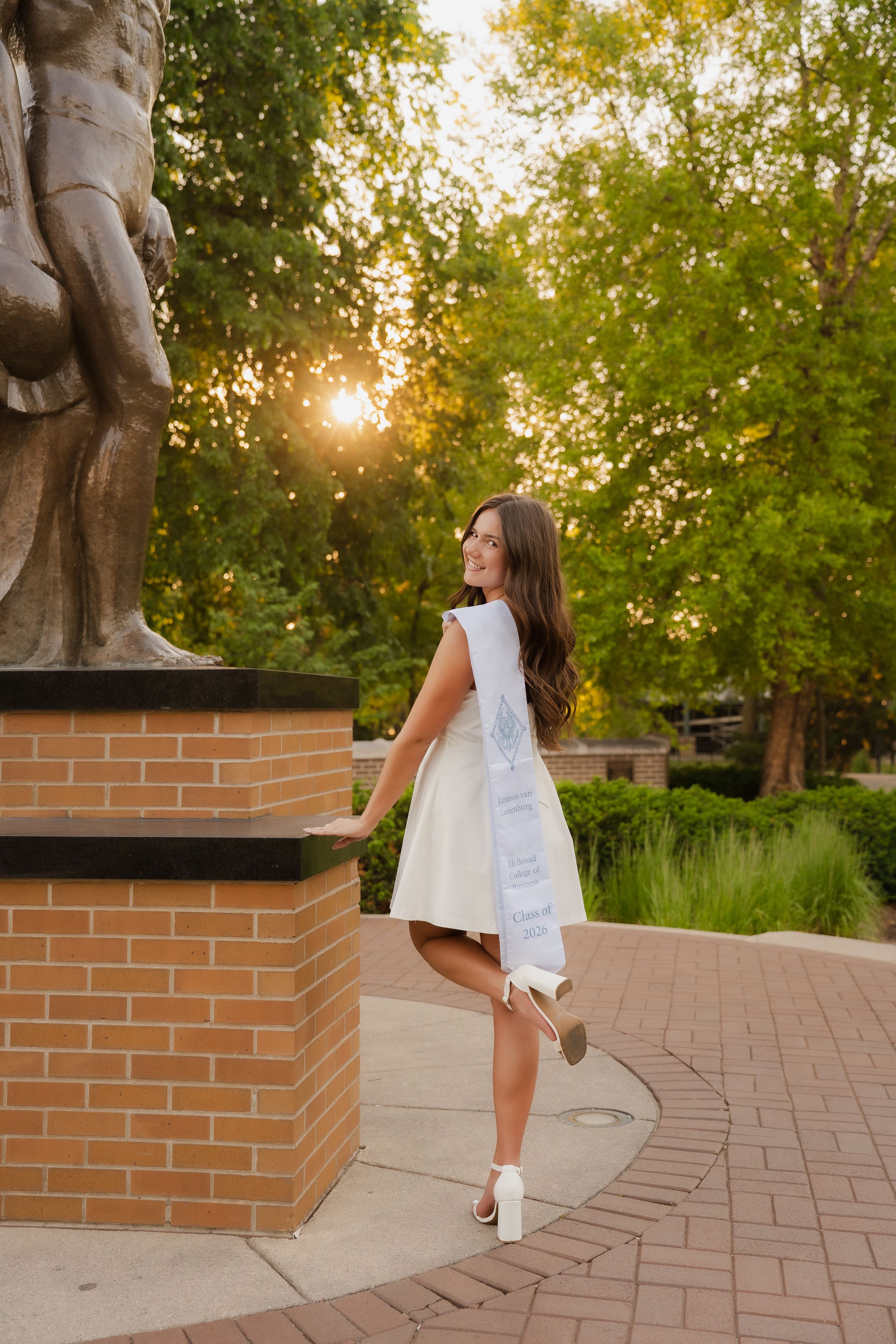 Young woman in white dress and graduation sash posing by brick statue pedestal outdoors at golden hour.