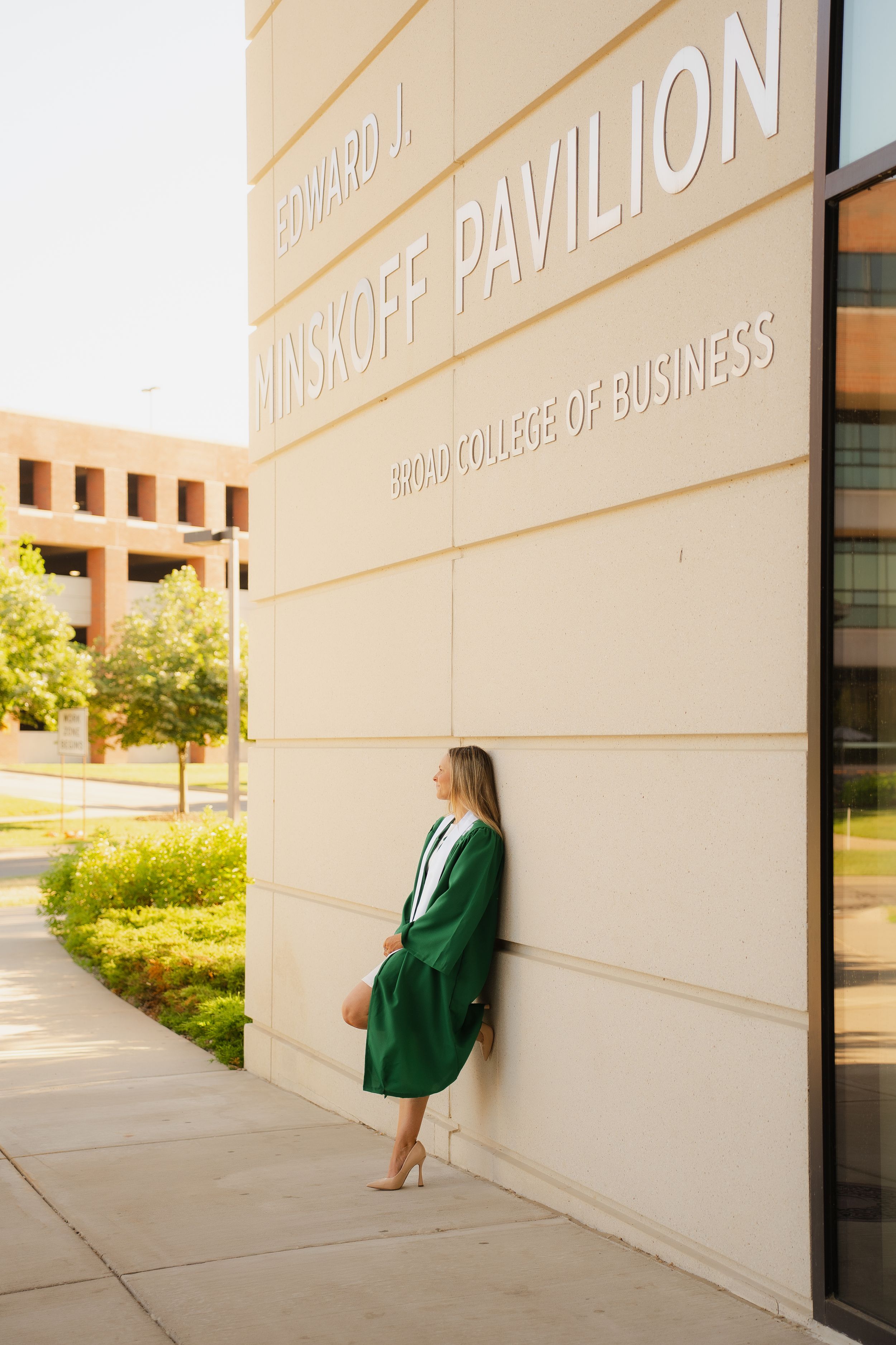 Graduate in green cap and gown posing at Edward J. Minskoff Pavilion, Harman College of Business.