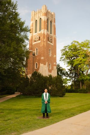Graduate in green gown stands before Beaumont Tower at Michigan State University on a sunny day.