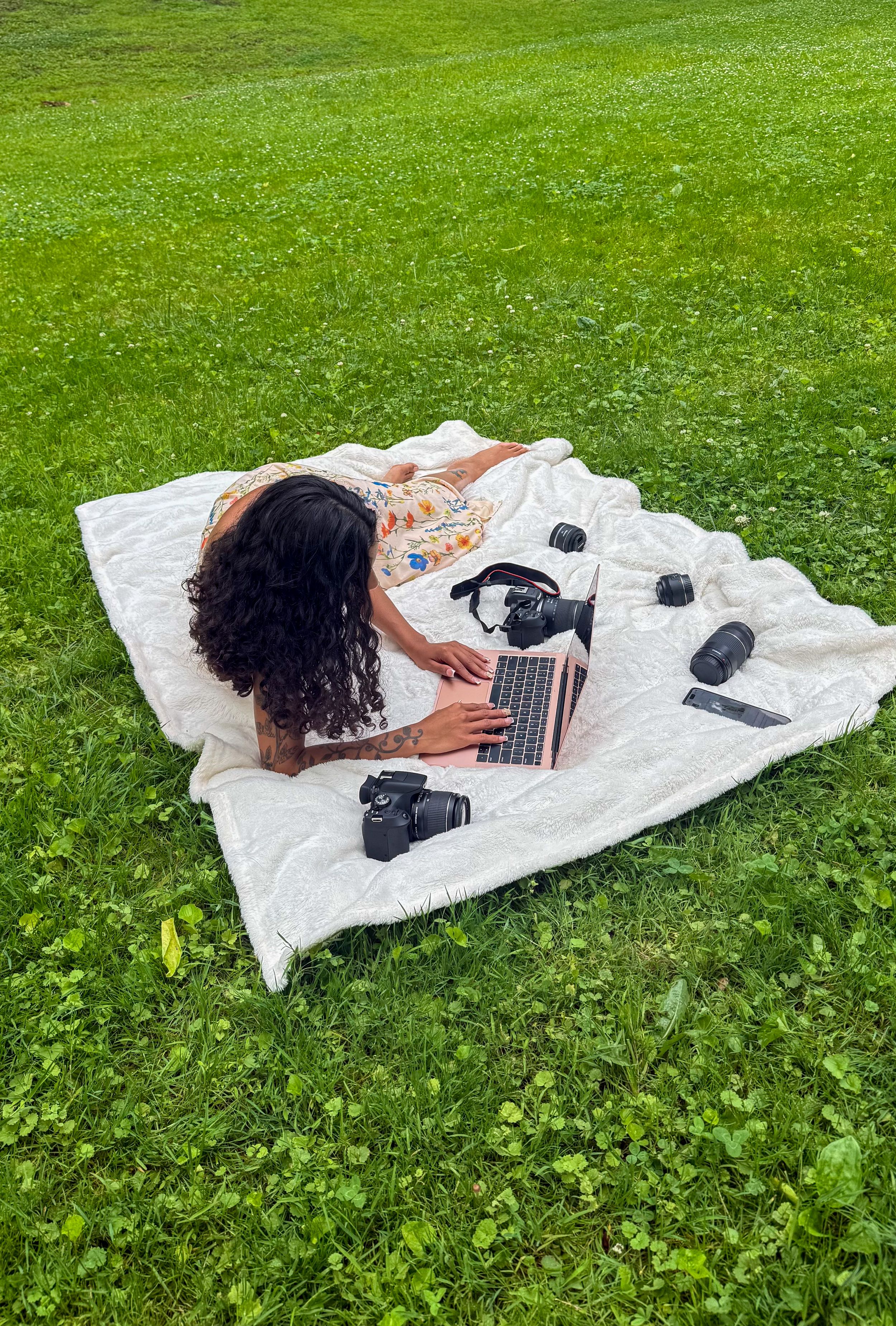 Woman with curly hair works on laptop outdoors on white blanket surrounded by DSLR cameras and headphones on green grass.