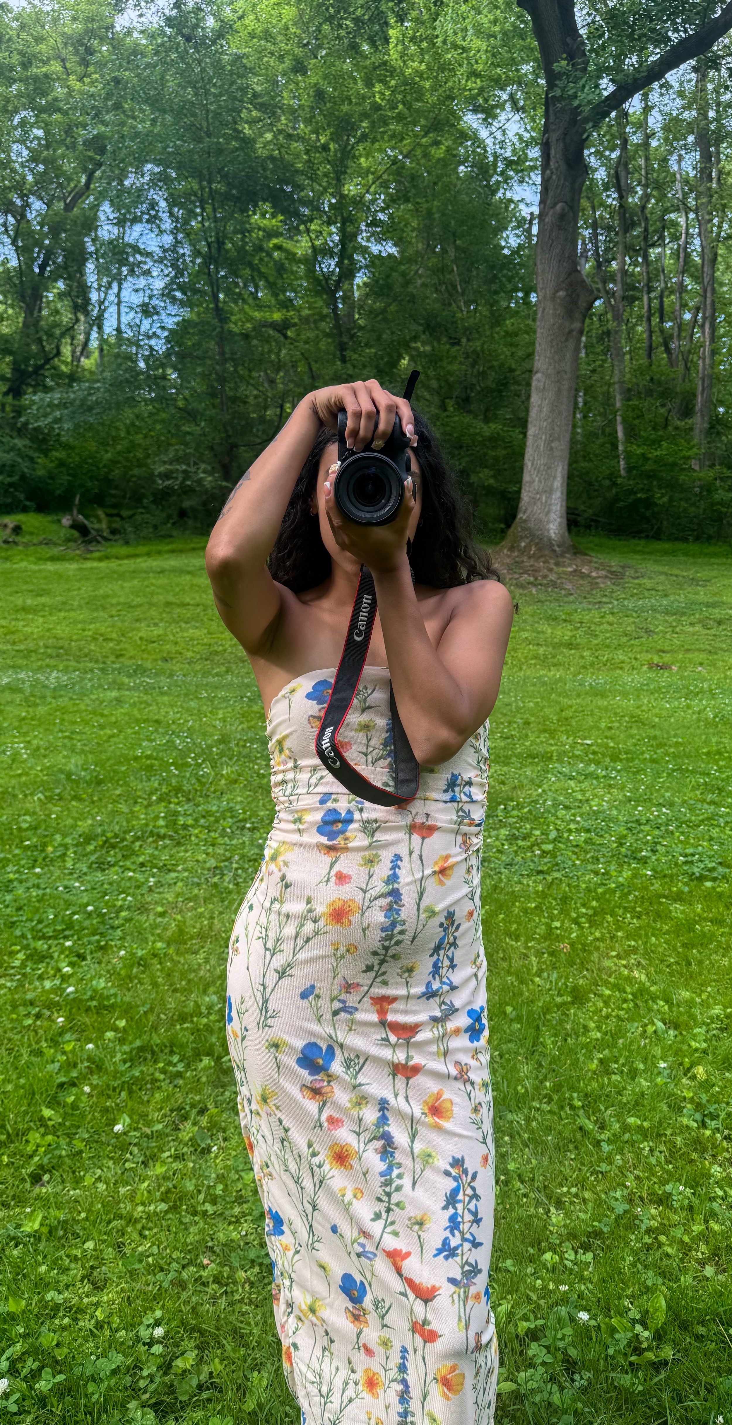Woman in floral maxi dress photographing with Canon camera outdoors in lush green park setting.