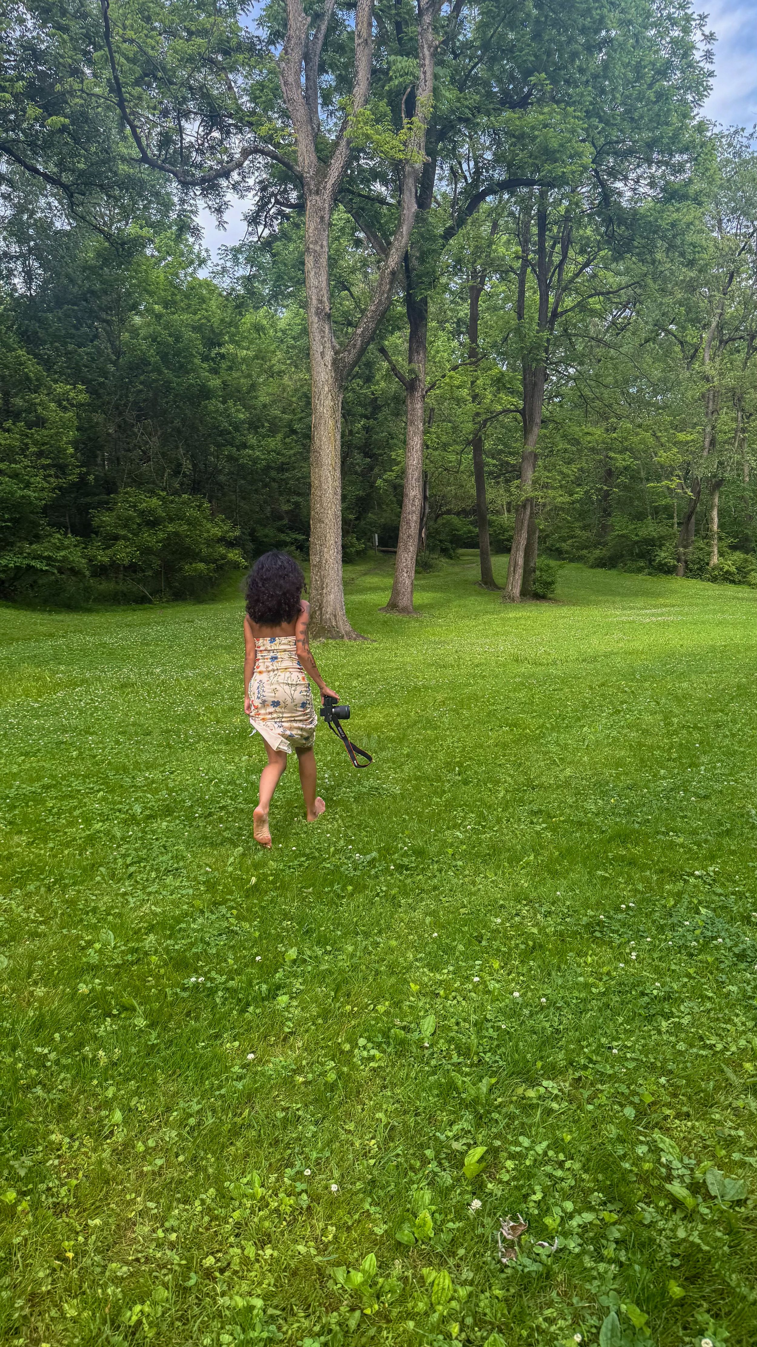A young girl in a floral dress runs barefoot across a lush green lawn toward a wooded area on a cloudy day.