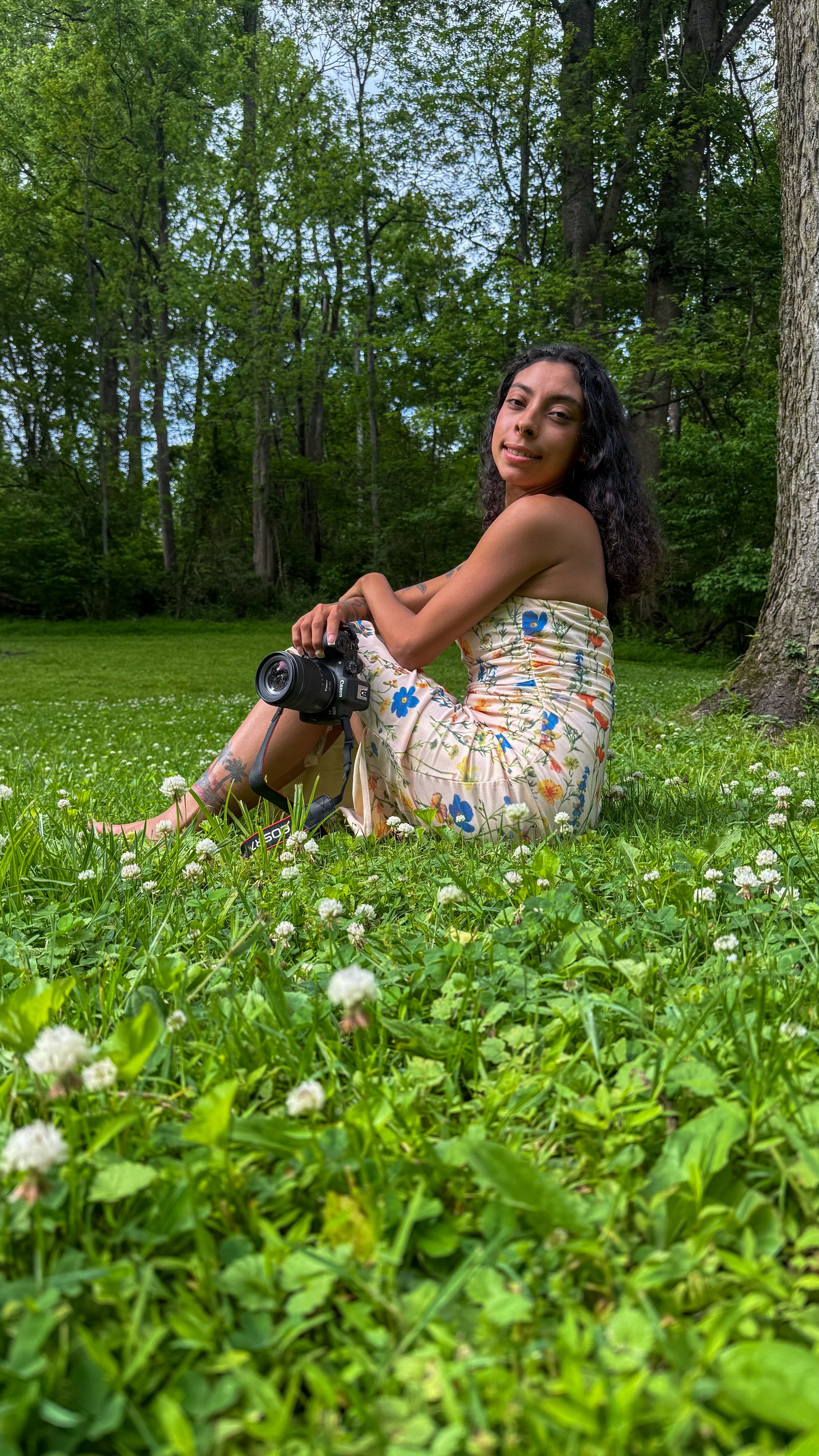Young woman in floral dress sitting on lush green grass holding a camera, surrounded by wildflowers and trees.