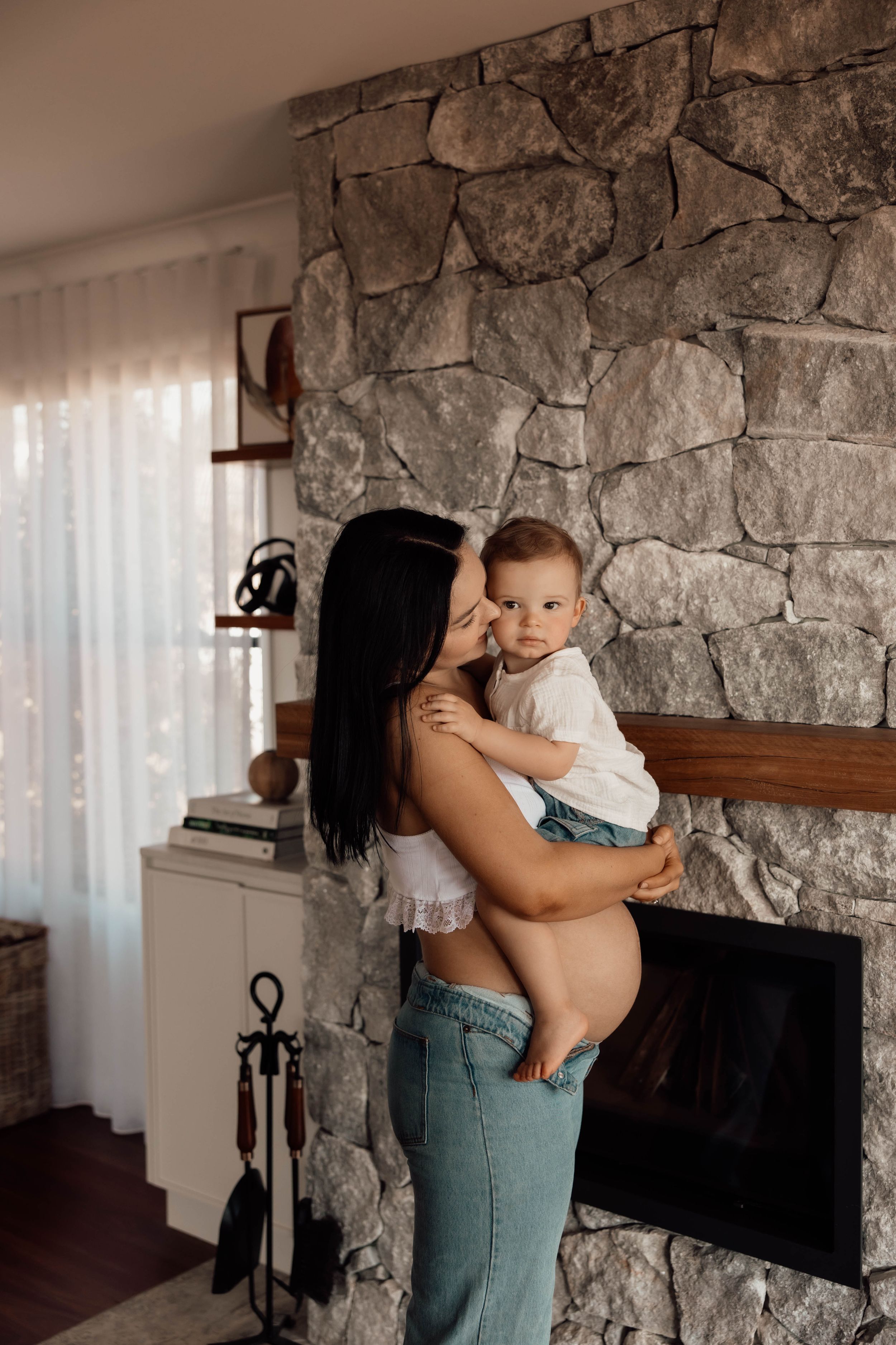 A young mother with dark hair holds her baby in front of a rustic stone fireplace in a cozy living room.