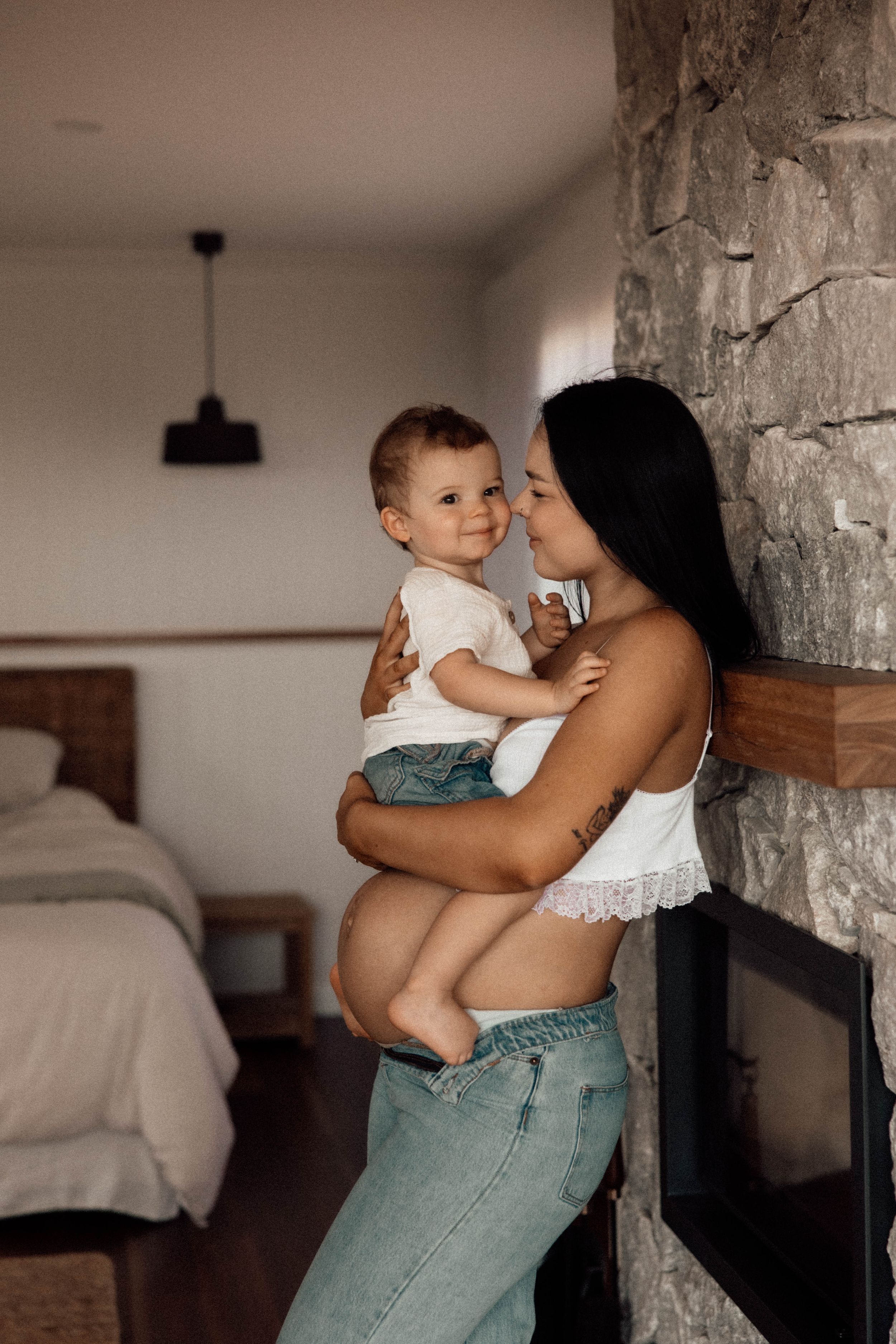 A young mother holds her smiling toddler while leaning against a stone fireplace in a modern bedroom.