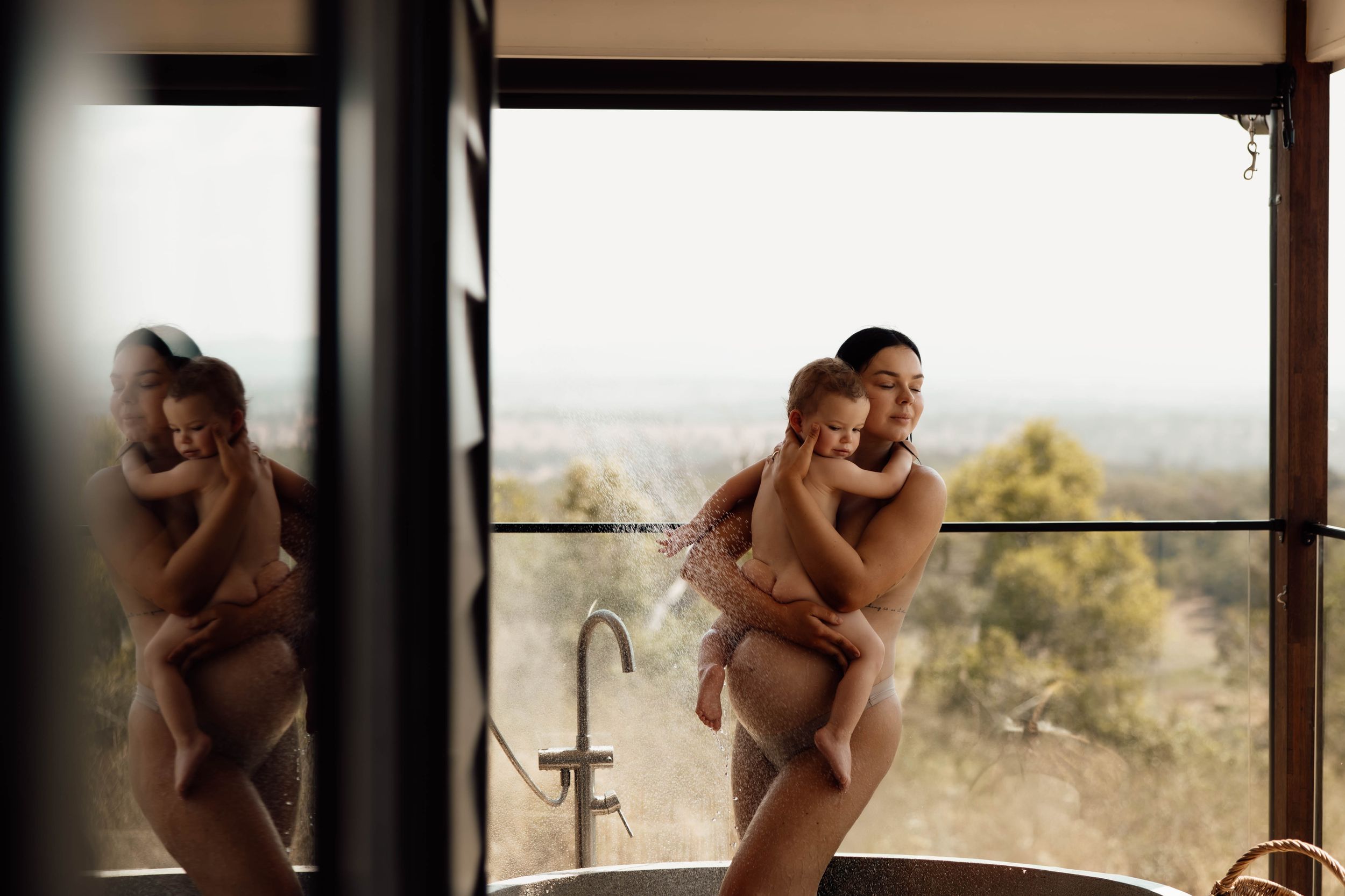 A mother tenderly holds her baby near a freestanding bathtub by a large window with scenic views.