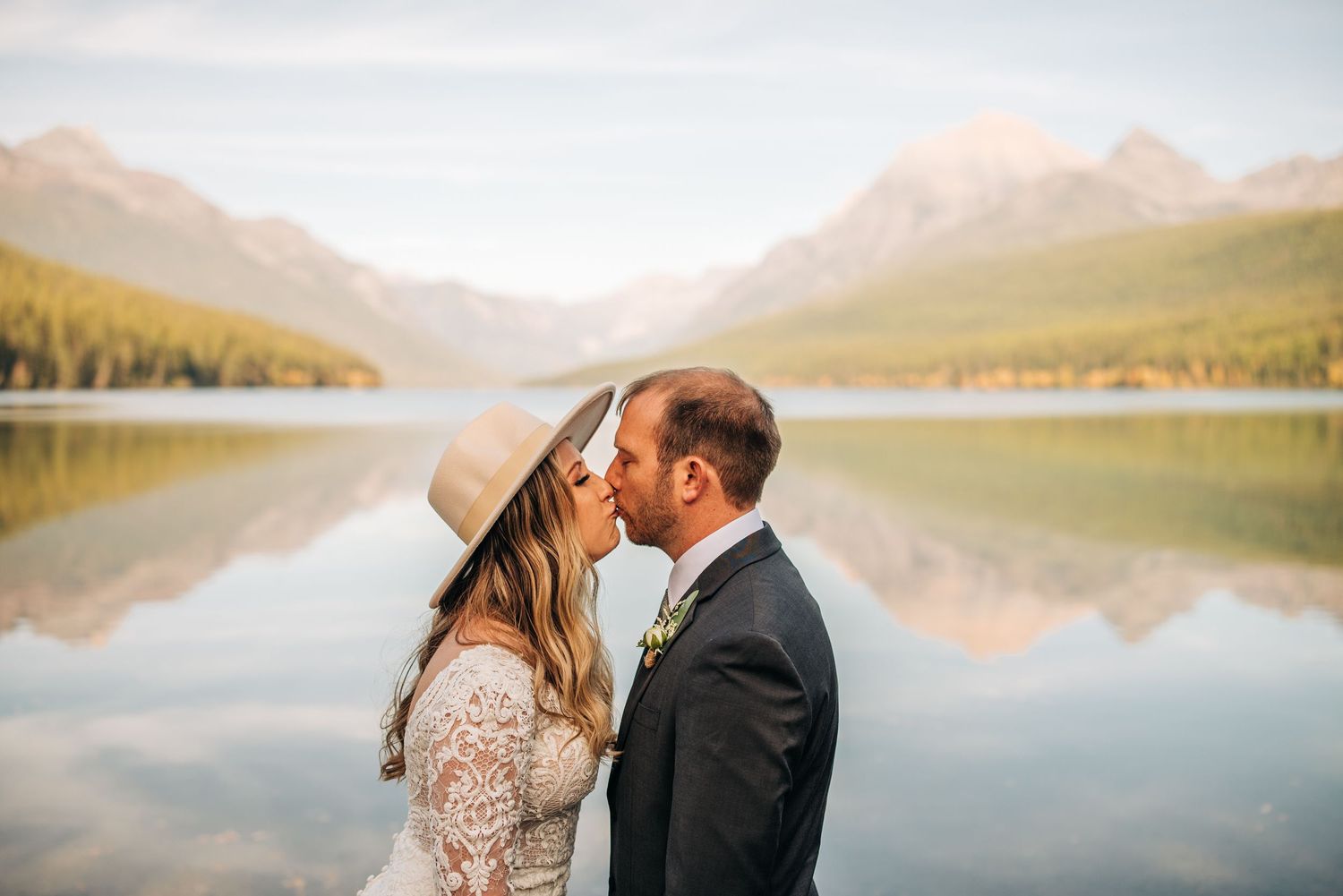 Couple sharing a kiss in front of a serene mountain lake reflection, bride wearing lace dress and tan hat.