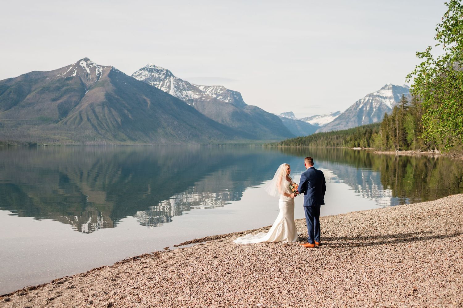 Bride and groom hold hands by a reflective mountain lake with snow-capped peaks in Glacier National Park.