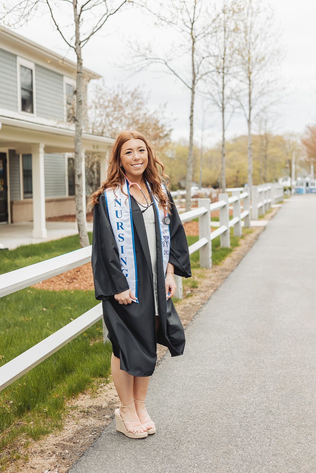 Smiling nursing graduate in black cap and gown with nursing sash poses on a tree-lined pathway near a white fence.