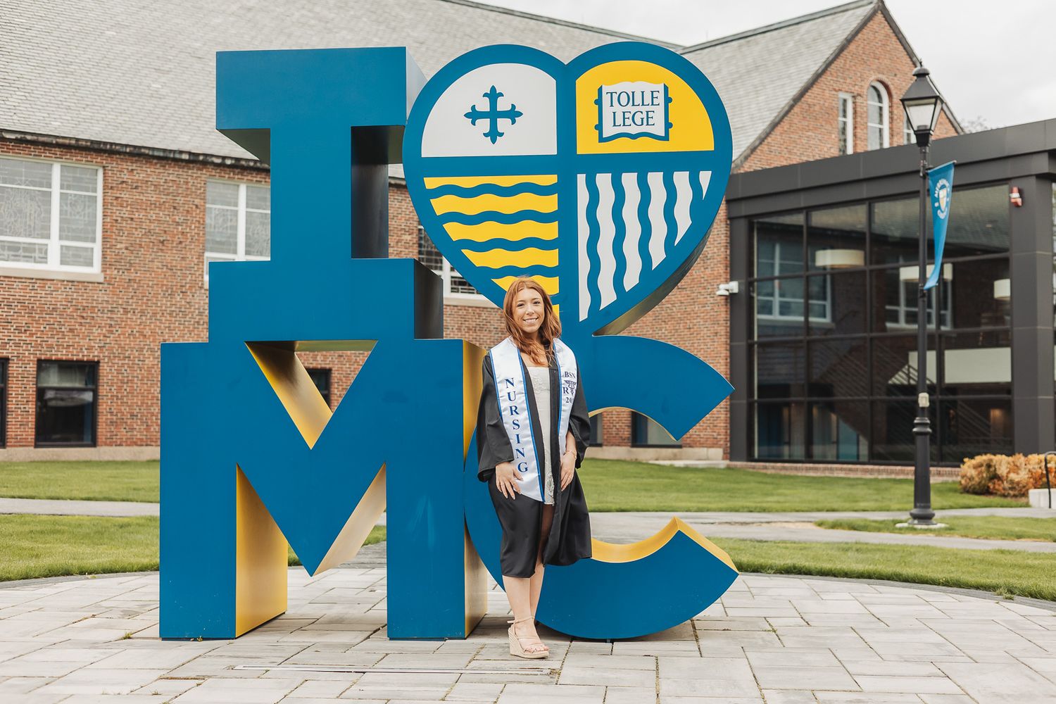 Graduate in cap and gown posing in front of a large blue I Love MC sign at Molloy College campus.