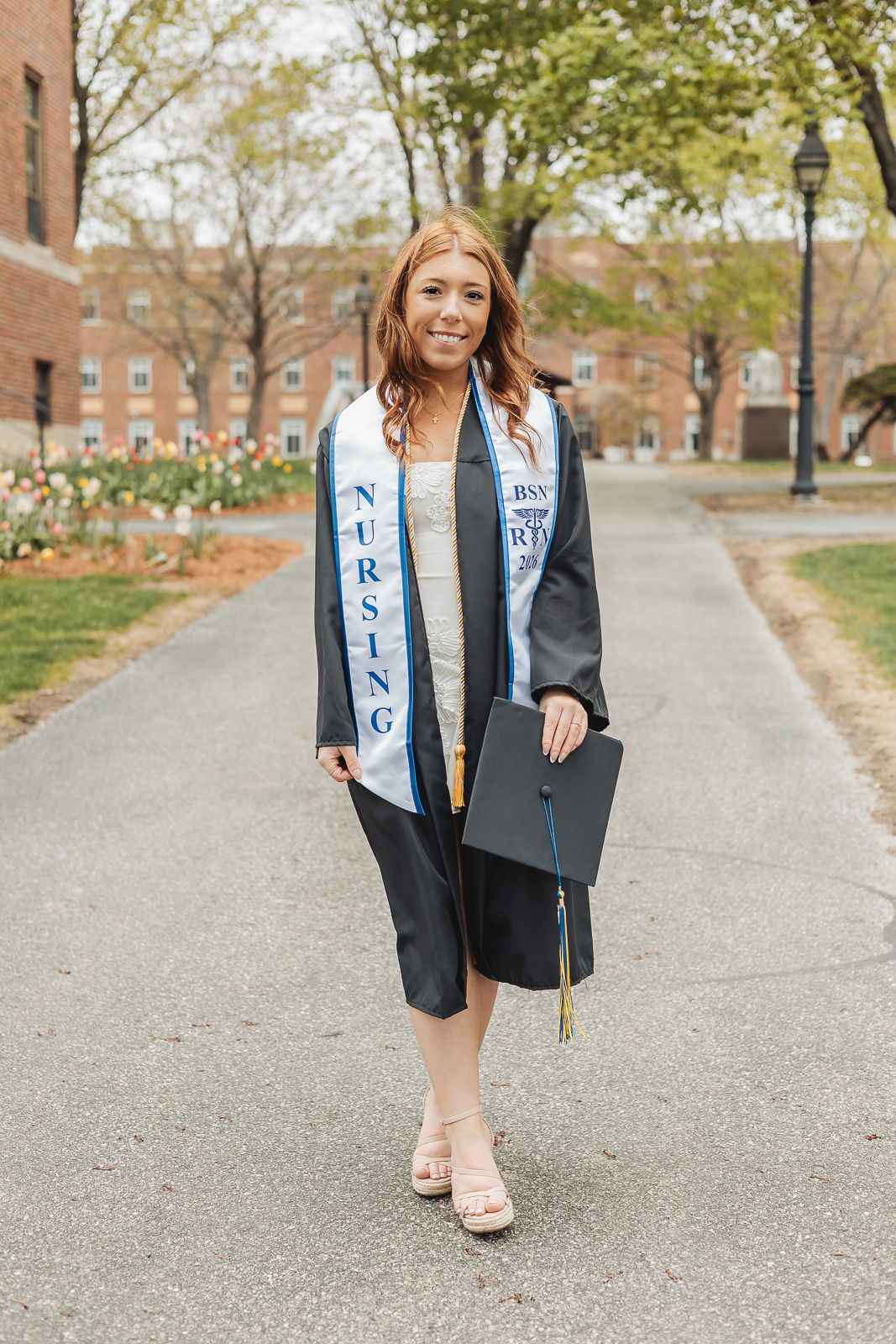 Nursing graduate in black cap and gown with BSN stole walks on campus path holding graduation cap.