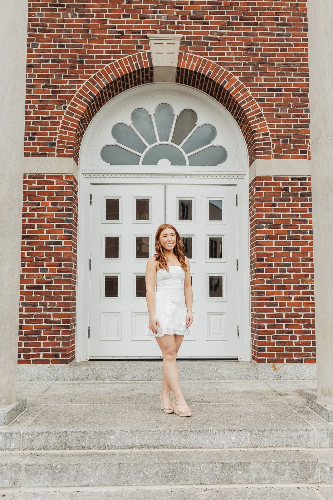 Young woman in white dress poses on steps before elegant white double doors framed by red brick arch.