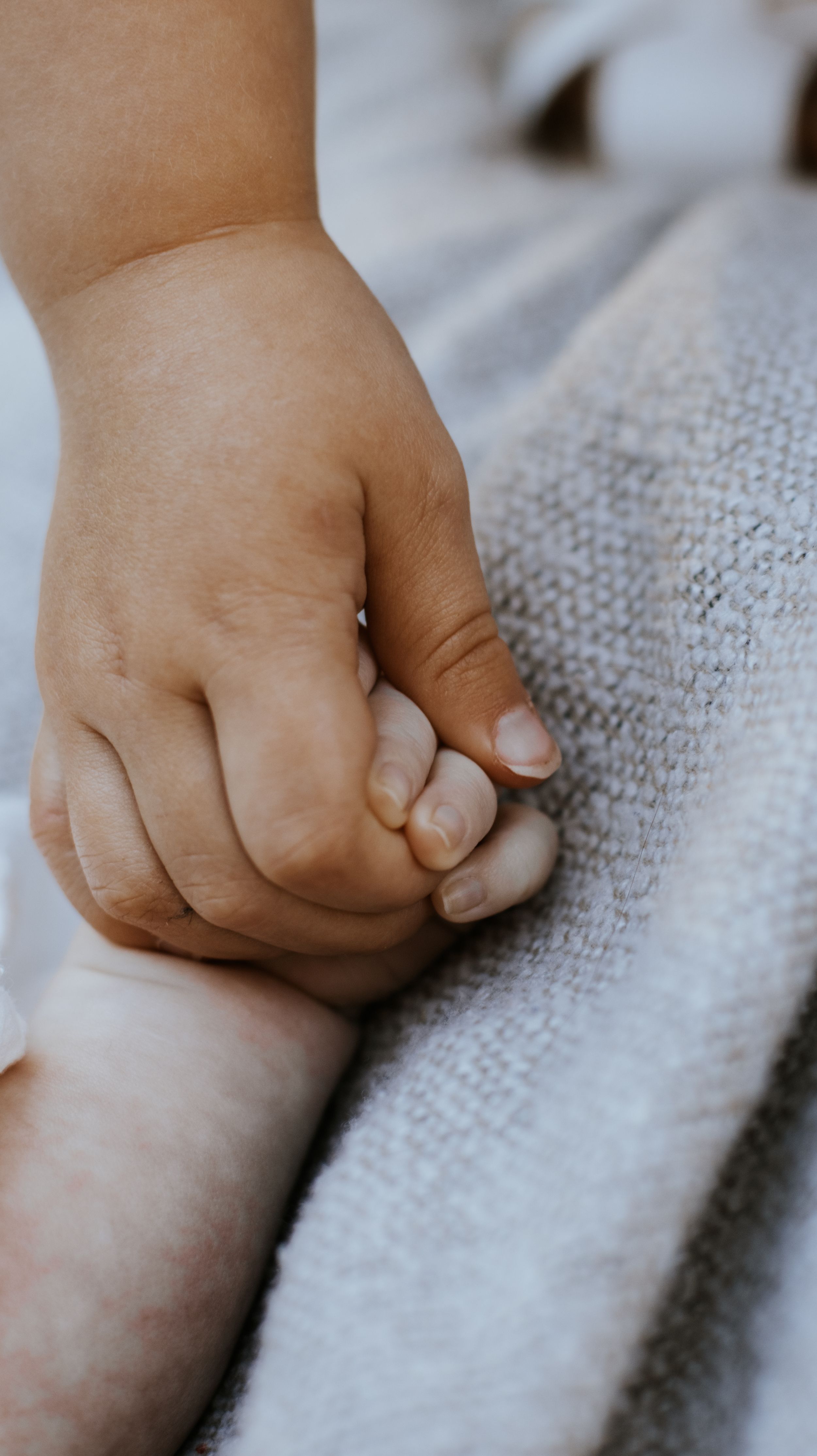 A tender close-up of a baby's tiny hand gently gripping an adult's finger on a soft knit blanket.
