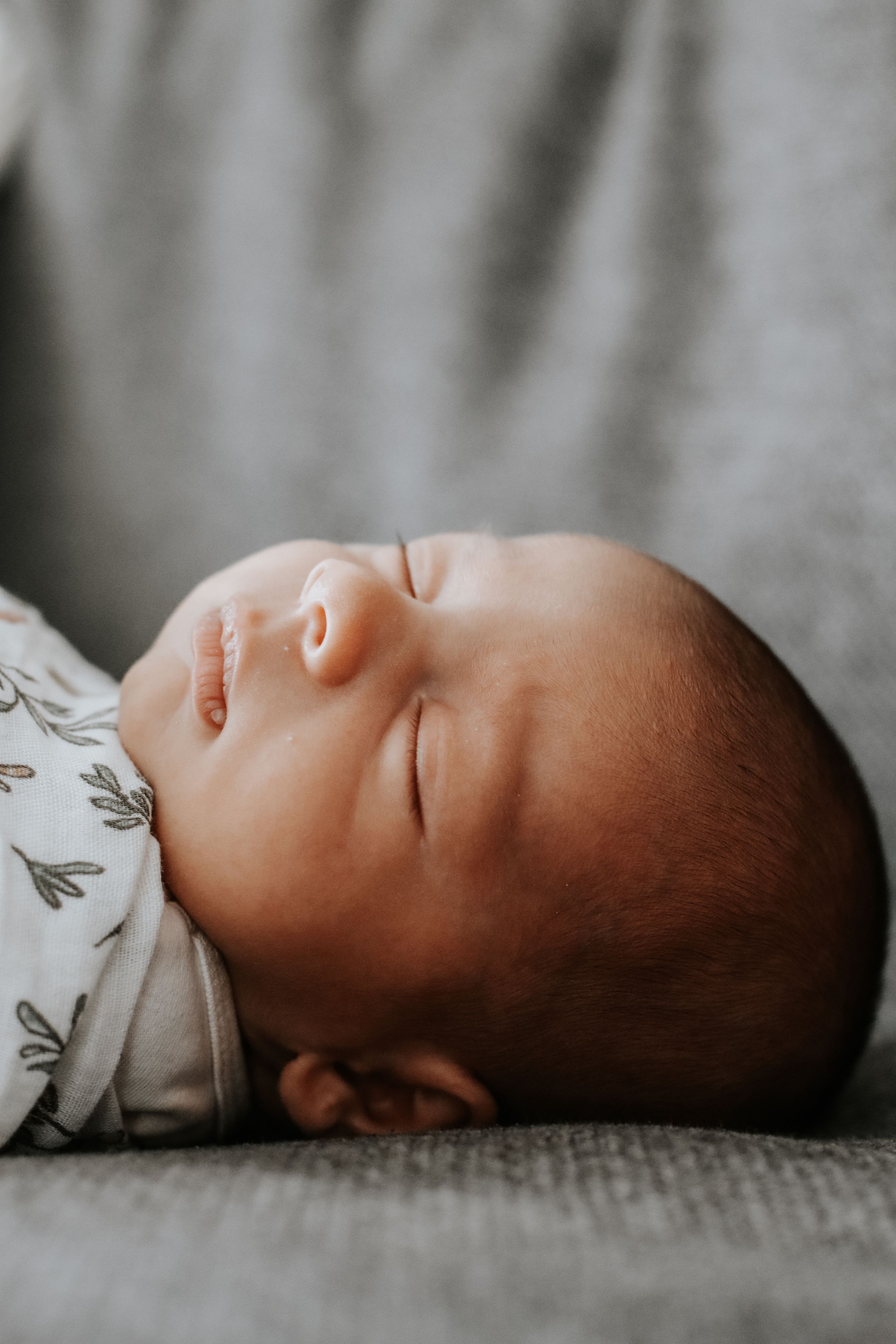 Peaceful newborn baby sleeping soundly, wearing a white floral onesie, lying on a soft gray blanket.