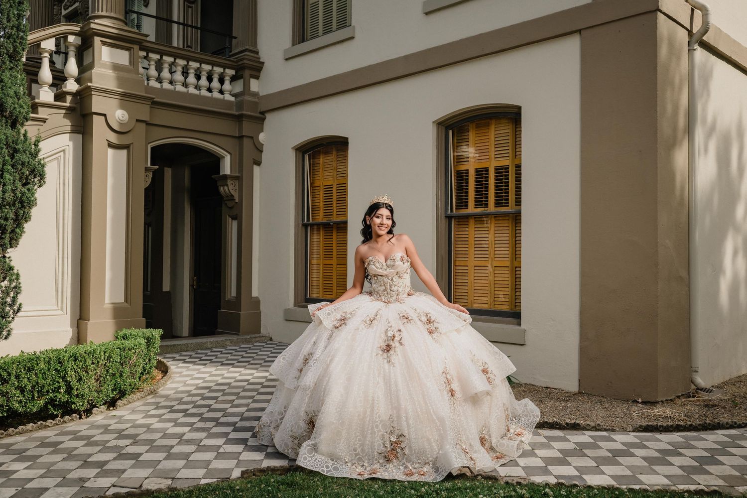 Bride in ballgown poses on lush lawn before a stunning white castle venue with Gothic arched windows and gold doors.