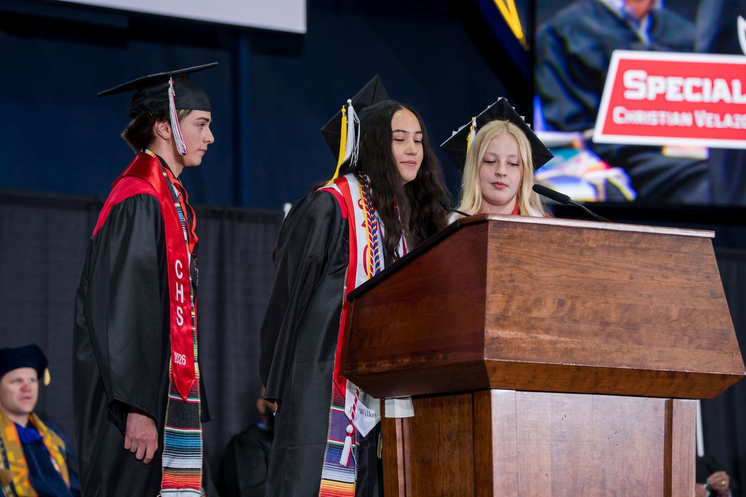 Two graduates in black caps and gowns stand at a podium during a graduation ceremony with a presenter nearby.
