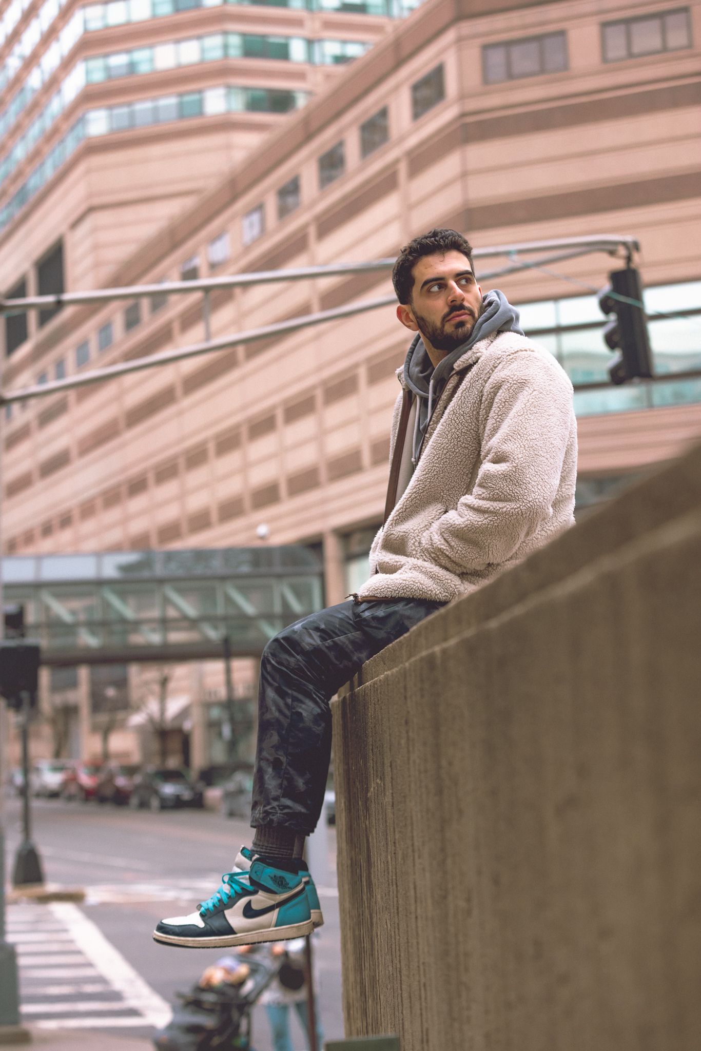 Young man in white sherpa hoodie, camo pants, and Nike Air Jordan sneakers sitting on urban concrete wall.