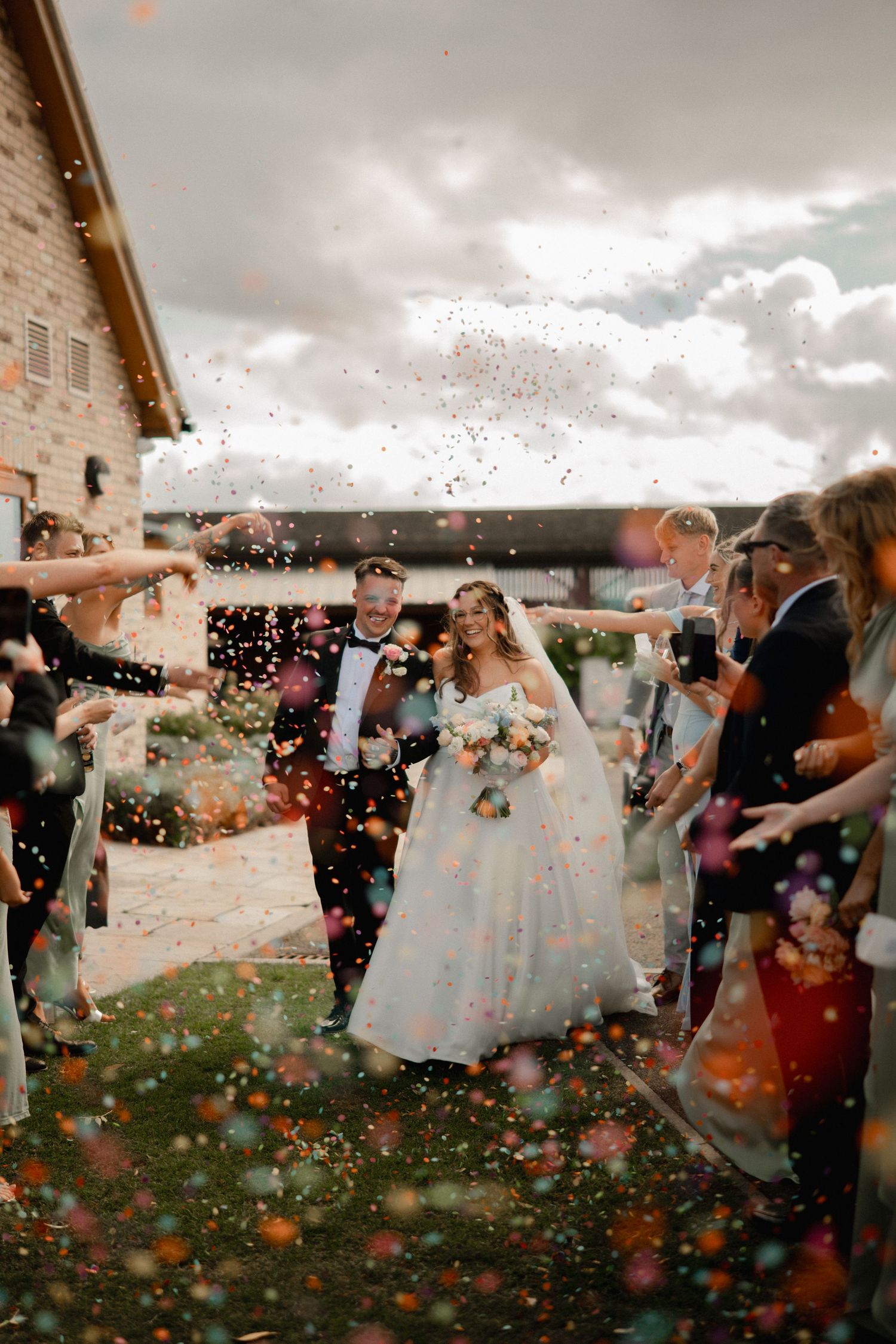 Bride and groom walk through colorful confetti shower surrounded by guests outside a rustic stone wedding venue.