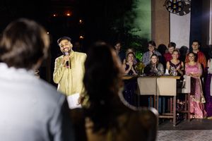 Man in gold suit speaking into microphone at elegant indoor celebration with guests in formal attire.