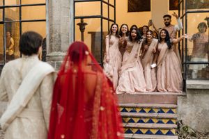 Bridesmaids in blush gowns cheer as Indian bride in red bridal veil approaches on ornate tiled steps.