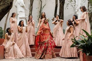 Bride in red lehenga surrounded by bridesmaids in pink lehengas celebrating on outdoor steps at Indian wedding.