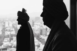 Two men in turbans silhouetted against a city skyline viewed from a high-rise window in black and white.