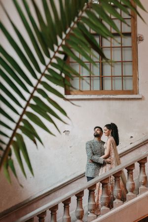 Couple in formal attire pose on white stone staircase framed by palm leaves.