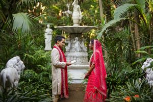 Couple in traditional Indian wedding attire poses by fountain in lush garden setting.