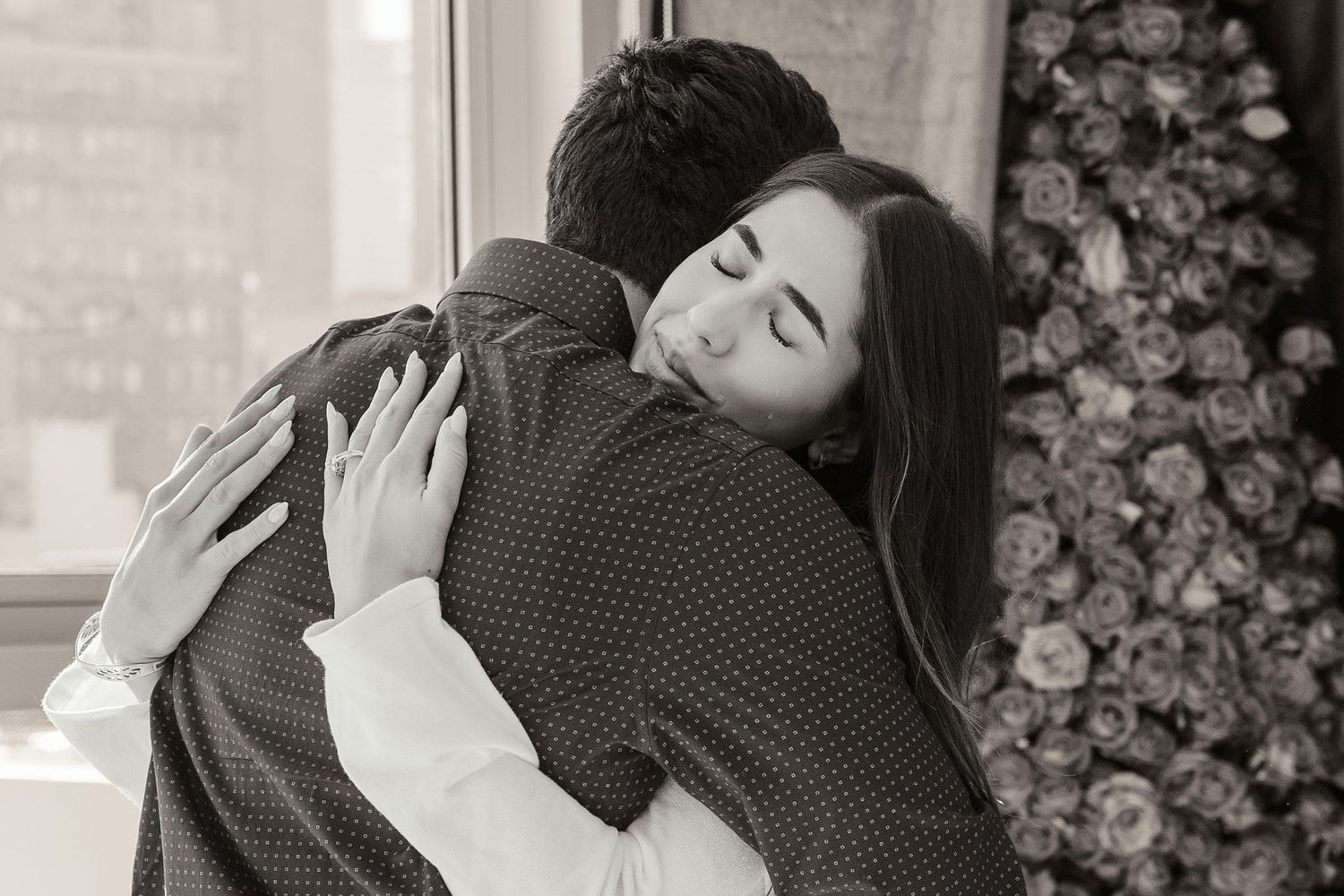 Black and white photo of a couple sharing a tender embrace near a window with floral decor in the background.