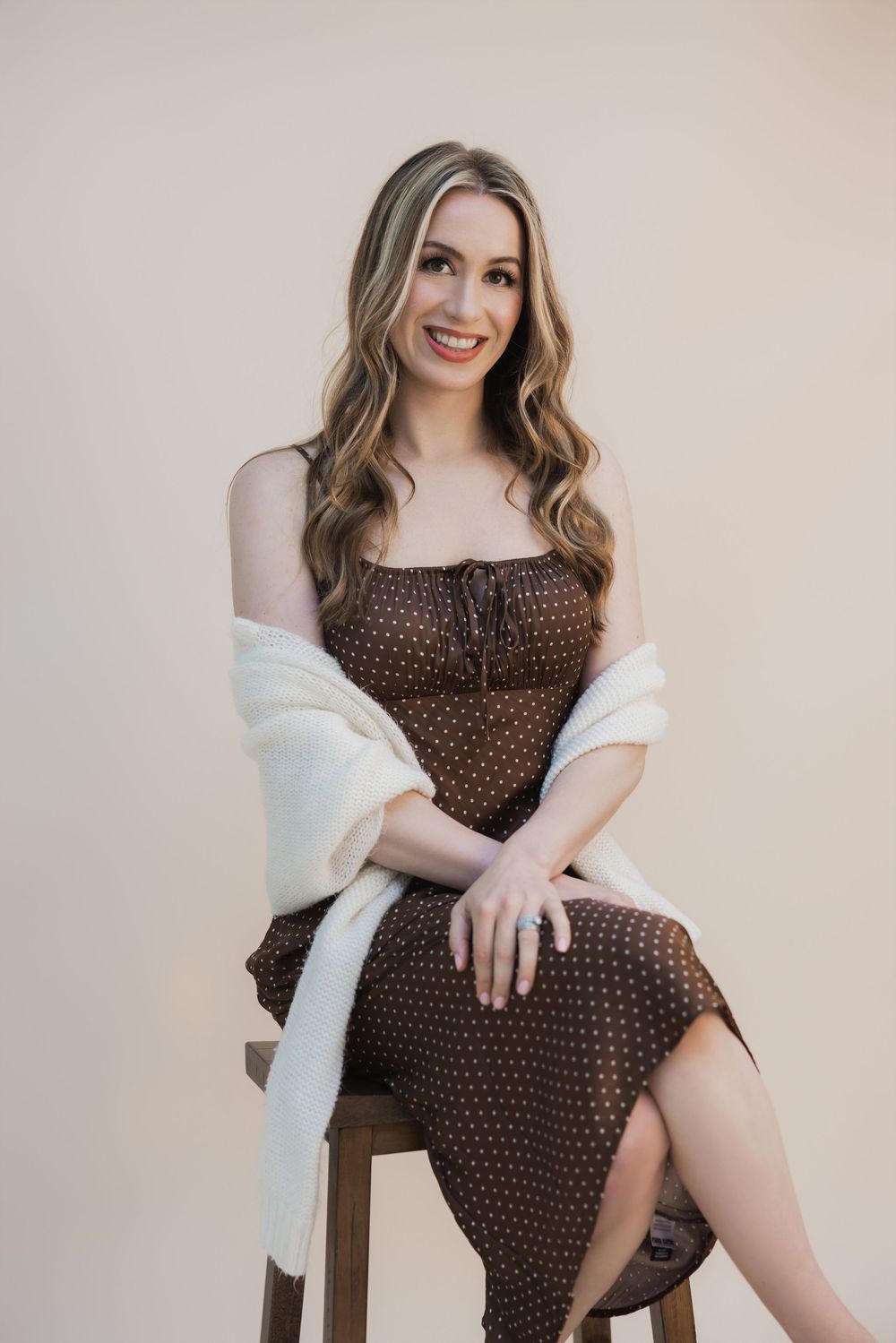 Woman in brown polka dot dress and white shawl sitting on wooden stool, smiling against beige background.