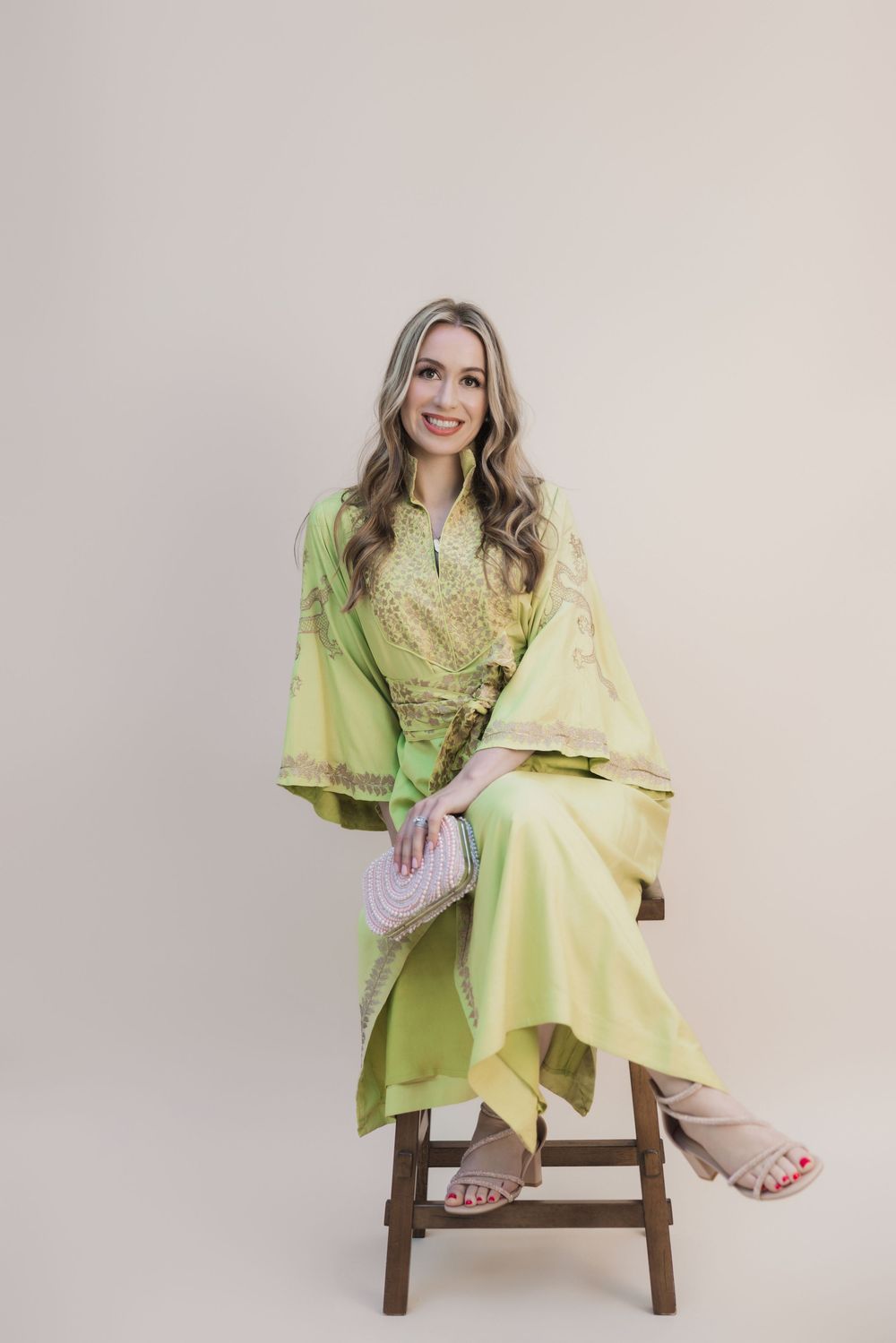 Woman sitting on wooden stool wearing lime green embroidered kaftan dress with wide sleeves, holding a small clutch purse.