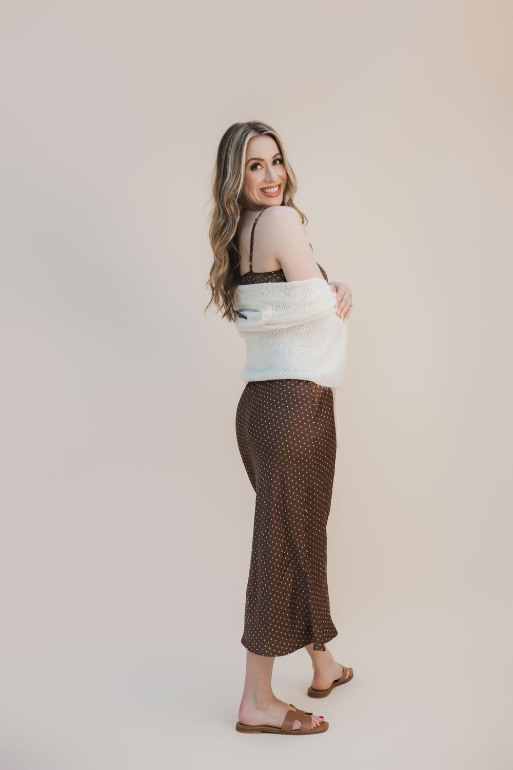 Woman posing in brown polka dot midi skirt with white fuzzy cardigan and tan slide sandals against neutral background.