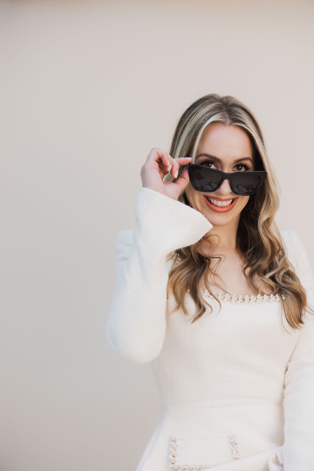 Smiling blonde woman in white outfit peeking over black sunglasses against a neutral background.