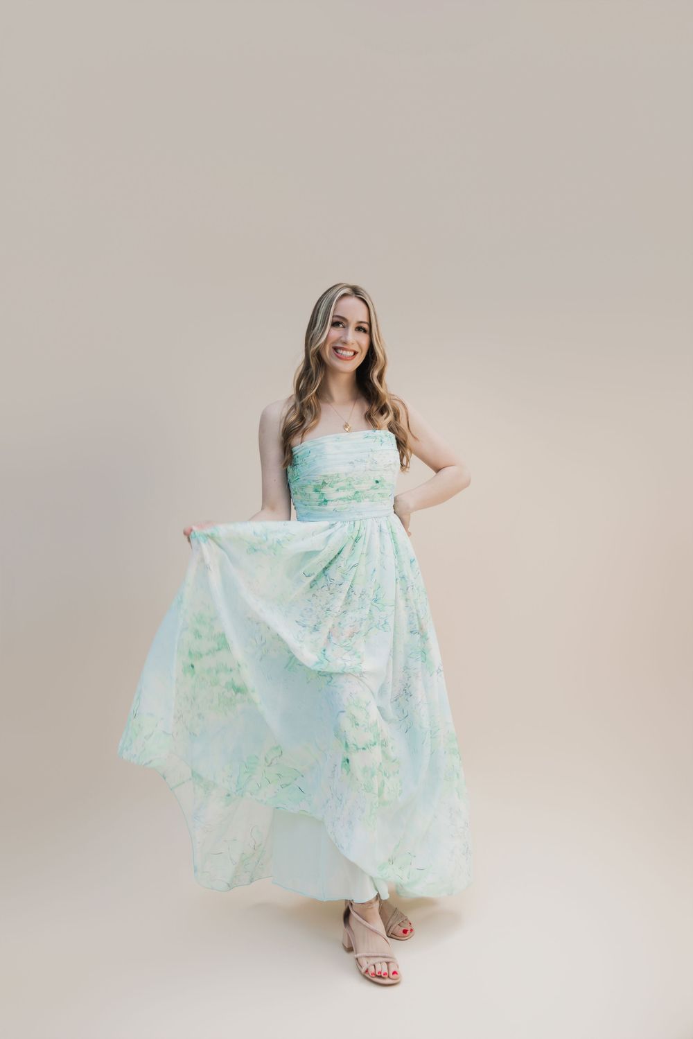 Woman in mint floral maxi dress posing gracefully, holding skirt, wearing nude heels on beige background.