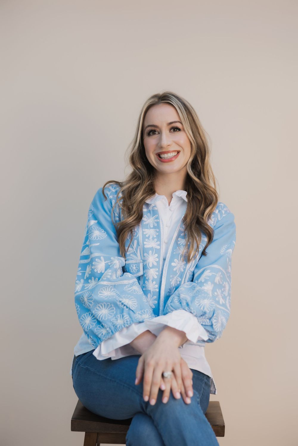 Smiling woman in blue embroidered blouse and jeans sitting on wooden stool against neutral background.