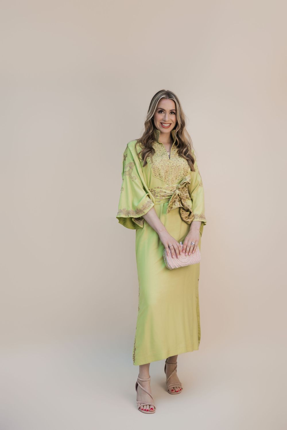 Woman in lime green kimono-style midi dress with floral embroidery, holding pink clutch, wearing nude heels.