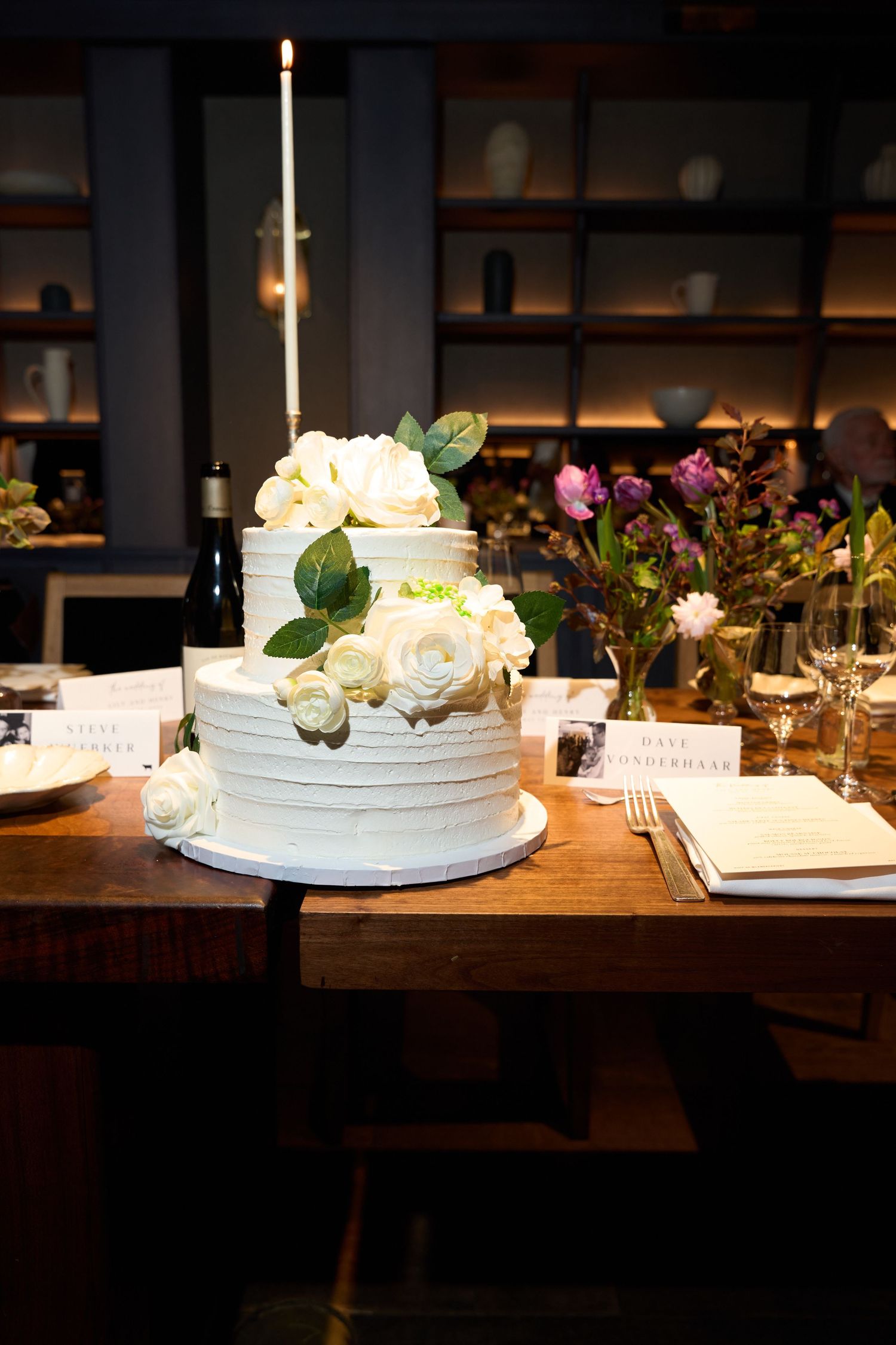 Elegant two-tier white wedding cake with textured frosting, adorned with white roses and green leaves.